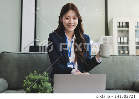 Young asian businesswoman eating a brownie while working from her office on a laptop next to a window with blinds Young asian businesswoman eating a brownie while working from her office on a laptop next to a window with blinds 141582721