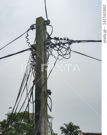 Close-up of a weathered concrete electricity pole with tangled chaotic power lines and ceramic insulators against an overcast sky Close-up of a weathered concrete electricity pole with tangled chaotic power lines and ceramic insulators against an overcast sky 141582902