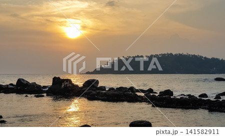 Sunset over rocky coastline with silhouetted tropical island and golden sun reflection on calm ocean, Mirissa, Sri Lanka Sunset over rocky coastline with silhouetted tropical island and golden sun reflection on calm ocean, Mirissa, Sri Lanka 141582911