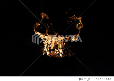 Bright burning wood fire inside a metal round grill bowl with glowing flames in the dark night. Close up view of outdoor campfire cooking setup with hot orange fire tongues shaping abstract patterns. Bright burning wood fire inside a metal round grill bowl with glowing flames in the dark night. Close up view of outdoor campfire cooking setup with hot orange fire tongues shaping abstract patterns. 141584532