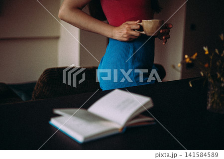 Woman holding a coffee cup at home with an open book, fresh flowers and a dog in the background. Cozy lifestyle and slow living comfort 141584589
