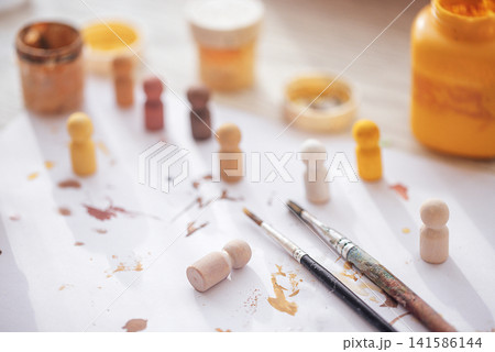 Wooden figurines and paintbrushes arranged on a white sheet with paint splatters, jars of paint in various colors visible in the background Wooden figurines and paintbrushes arranged on a white sheet with paint splatters, jars of paint in various colors visible in the background 141586144