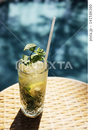 Refreshing mojito cocktail with mint leaves and lime served in a tall glass on a woven table beside a swimming pool under bright sunlight Refreshing mojito cocktail with mint leaves and lime served in a tall glass on a woven table beside a swimming pool under bright sunlight 141586160