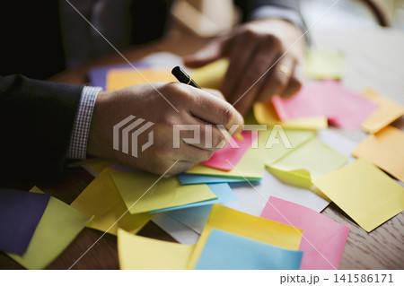Male office worker in a suit writing on a sticky note surrounded by colorful notes on a wooden desk, emphasizing organization and brainstorming activities Male office worker in a suit writing on a sticky note surrounded by colorful notes on a wooden desk, emphasizing organization and brainstorming activities 141586171