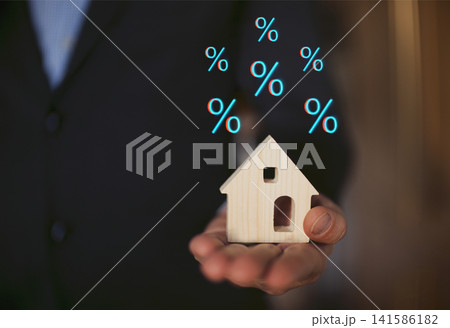 Businessman in a suit holds a small wooden house model in one hand with floating percentage symbols above, representing real estate investment and mortgage rates Businessman in a suit holds a small wooden house model in one hand with floating percentage symbols above, representing real estate investment and mortgage rates 141586182