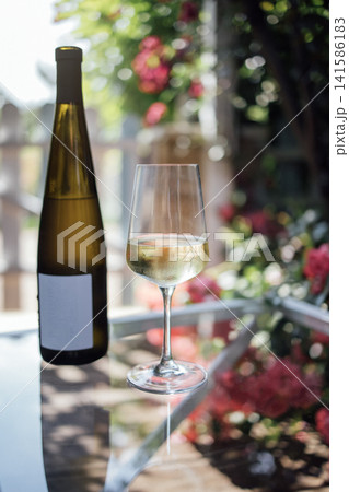White wine bottle and glass filled with white wine placed on a glass table surrounded by blooming flowers in a sunny outdoor setting White wine bottle and glass filled with white wine placed on a glass table surrounded by blooming flowers in a sunny outdoor setting 141586183