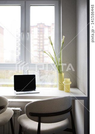 Modern workspace featuring a laptop on a white desk beside a window with a view of buildings, complemented by a yellow vase and decorative plants 141586448