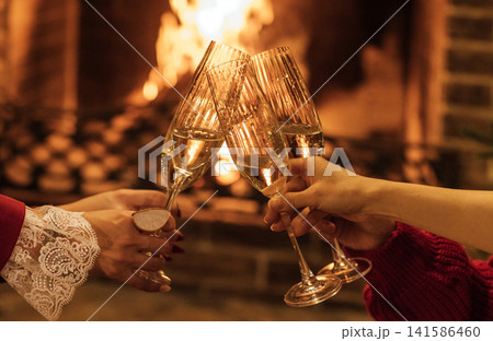 Hands of two women clinking champagne glasses in front of a cozy fireplace, warm glow illuminating the scene with festive atmosphere and holiday spirit Hands of two women clinking champagne glasses in front of a cozy fireplace, warm glow illuminating the scene with festive atmosphere and holiday spirit 141586460