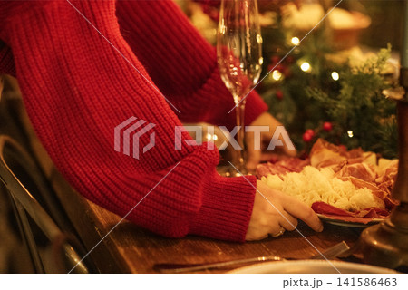 Hands of a person in a red sweater arranging a festive charcuterie board with assorted meats and cheeses on a wooden table with holiday decorations and glassware 141586463