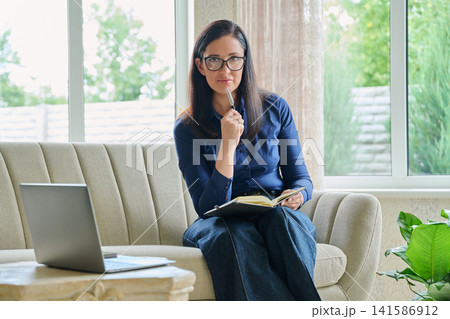 Serious businesswoman writing in notebook looking at camera at modern home office 141586912