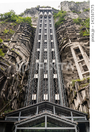 A towering multi-shaft glass elevator climbing vertically against a majestic, weathered karst cliff face, bridging the gap between earth and sky. 141587575