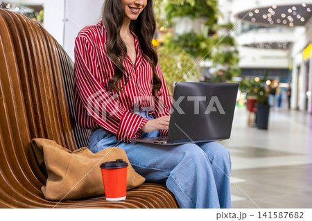 Young woman with a laptop sitting on the bench in a shopping mall 141587682