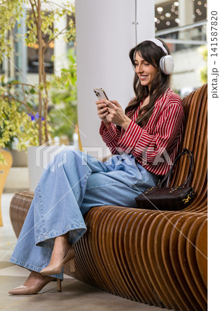 Young woman sitting on a bench in a shopping mall and listening to music Young woman sitting on a bench in a shopping mall and listening to music 141587820