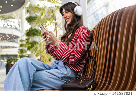 Young woman sitting on a bench in a shopping mall and listening to music Young woman sitting on a bench in a shopping mall and listening to music 141587821
