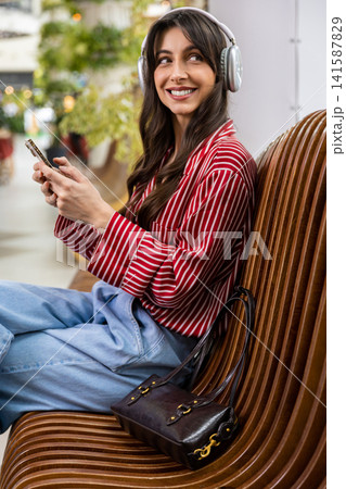 Young woman sitting on a bench in a shopping mall and listening to music Young woman sitting on a bench in a shopping mall and listening to music 141587829