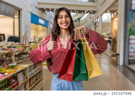 Smiling young woman feeling happy and contented while doing shopping in a shopping center Smiling young woman feeling happy and contented while doing shopping in a shopping center 141587946