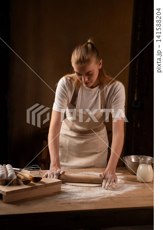Woman baking fresh bread or pastry dough on rustic wooden kitchen table, rolling it with pin, creating artisan food Woman baking fresh bread or pastry dough on rustic wooden kitchen table, rolling it with pin, creating artisan food 141588204
