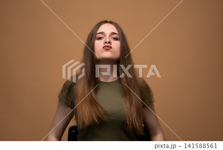 Young woman showing playful attitude with a pouty face on a brown background, expressing disapproval or stubbornness Young woman showing playful attitude with a pouty face on a brown background, expressing disapproval or stubbornness 141588561