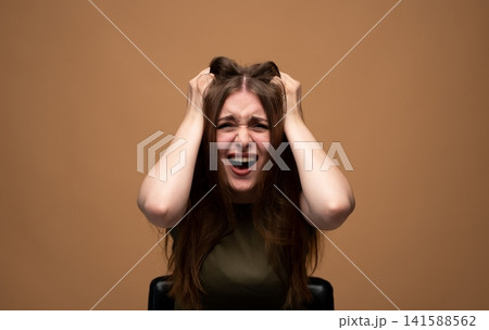 Young woman suffering from intense stress and frustration, screaming in anger while pulling her long hair Young woman suffering from intense stress and frustration, screaming in anger while pulling her long hair 141588562