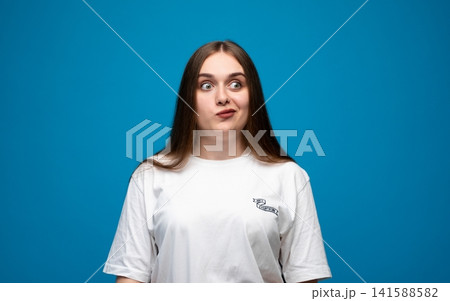 Young woman having an unsure and curious facial expression, looking away with doubt, standing against a blue background 141588582