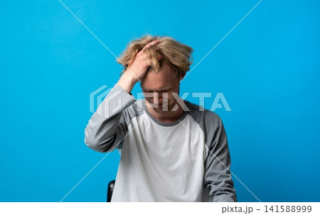 Young man experiencing severe headache, stress, and frustration, grimacing and holding his head in discomfort, suffering from pain and mental distress on blue background Young man experiencing severe headache, stress, and frustration, grimacing and holding his head in discomfort, suffering from pain and mental distress on blue background 141588999