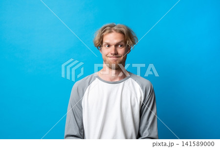 Young adult man making a skeptical expression with a smirk, feeling doubtful, suspicious, and uncertain, looking away on a blue background 141589000