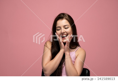 Happy young woman expressing joy and surprise with hands on cheeks, smiling broadly on a pink background, feeling excited Happy young woman expressing joy and surprise with hands on cheeks, smiling broadly on a pink background, feeling excited 141589013