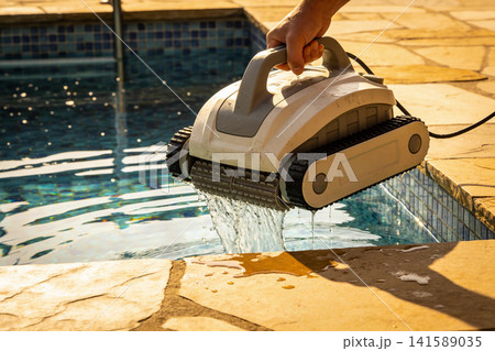Hand holding robotic pool cleaner with water dripping into swimming pool Hand holding robotic pool cleaner with water dripping into swimming pool 141589035