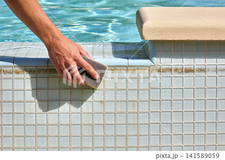 Hand scrubbing waterline tiles in a swimming pool with a cleaning sponge 141589059