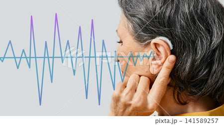 Hearing aids. Senior woman wearing behind the ear hearing aid with sound waves pattern in foreground, concept deafness treatment Hearing aids. Senior woman wearing behind the ear hearing aid with sound waves pattern in foreground, concept deafness treatment 141589257