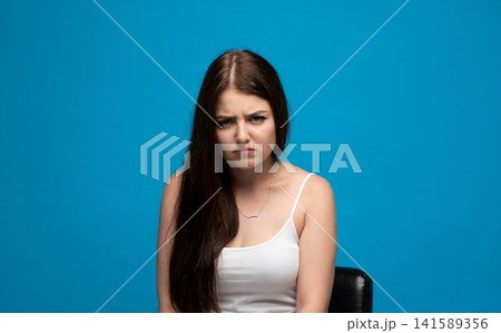 Young woman showing frustration and anger, having a displeased expression against a vibrant blue background Young woman showing frustration and anger, having a displeased expression against a vibrant blue background 141589356