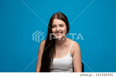 Young woman smiling, looking at camera with joy and happiness on blue background, celebrating success Young woman smiling, looking at camera with joy and happiness on blue background, celebrating success 141589395