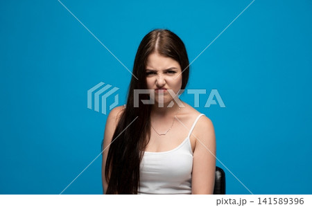 Young woman showing strong disgust, grimacing with a displeased and upset facial expression on blue background Young woman showing strong disgust, grimacing with a displeased and upset facial expression on blue background 141589396