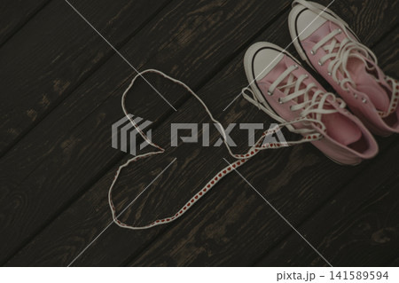 Pastel pink sneakers with shoelaces arranged in a heart shape on a dark wooden background. Romance, authenticity, and modern relationships concepts. Pastel pink sneakers with shoelaces arranged in a heart shape on a dark wooden background. Romance, authenticity, and modern relationships concepts. 141589594
