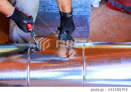 Worker uses snips to cut metal circle from duct it is tools are visible nearby. 141589703