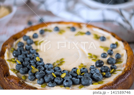 Round dessert features cream cheese filling topped with fresh blueberries, pumpkin seeds on wooden table. Round dessert features cream cheese filling topped with fresh blueberries, pumpkin seeds on wooden table. 141589746