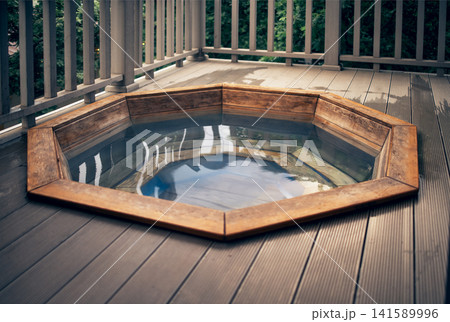 Wooden octagonal hot tub filled with water on a wooden deck surrounded by a railing, reflecting trees and sky in the water surface 141589996