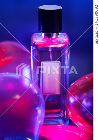 Glass perfume bottle with a black cap surrounded by colorful balloons illuminated by vibrant pink and blue lighting on a blurred background Glass perfume bottle with a black cap surrounded by colorful balloons illuminated by vibrant pink and blue lighting on a blurred background 141590092