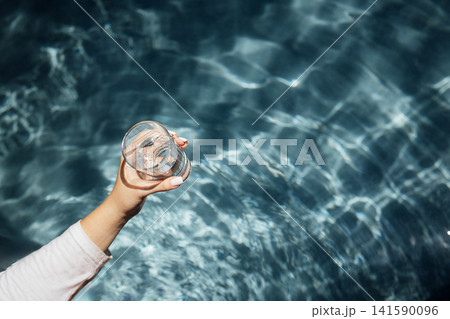 Hand holding a clear glass of water above shimmering blue water surface, sunlight reflecting off the water creating a sparkling effect Hand holding a clear glass of water above shimmering blue water surface, sunlight reflecting off the water creating a sparkling effect 141590096