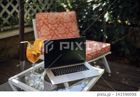 Outdoor workspace featuring a laptop on a glass table beside a refreshing cocktail in a garden setting with a patterned chair and lush greenery Outdoor workspace featuring a laptop on a glass table beside a refreshing cocktail in a garden setting with a patterned chair and lush greenery 141590098