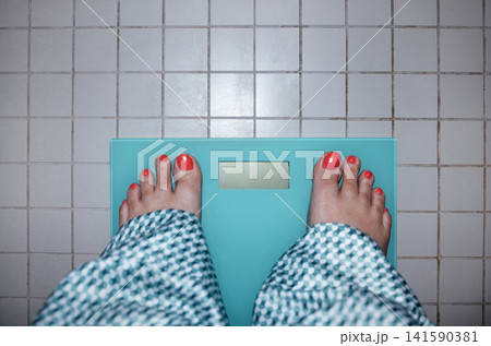 Bare feet with bright red nail polish standing on a turquoise bathroom scale on tiled floor, checkered bathrobe visible around ankles in a domestic setting Bare feet with bright red nail polish standing on a turquoise bathroom scale on tiled floor, checkered bathrobe visible around ankles in a domestic setting 141590381