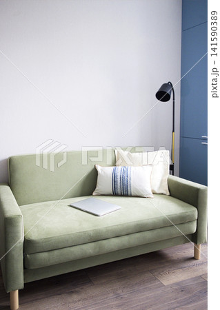 Green fabric sofa with decorative pillows and a laptop on it, positioned against a light wall and a blue cabinet with a standing lamp nearby 141590389