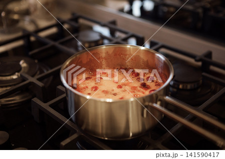 Stainless steel pot with bubbling red sauce containing tomatoes on a gas stove, surrounded by burners and kitchen appliances in a modern kitchen setting Stainless steel pot with bubbling red sauce containing tomatoes on a gas stove, surrounded by burners and kitchen appliances in a modern kitchen setting 141590417