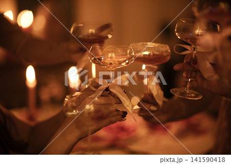 Group of hands holding champagne glasses with ribbons, celebrating at a candlelit table adorned with soft lighting and blurred background elements Group of hands holding champagne glasses with ribbons, celebrating at a candlelit table adorned with soft lighting and blurred background elements 141590418