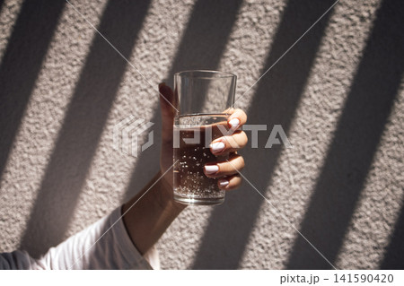 Hand holding a clear glass of sparkling water with bubbles, sunlight casting shadows on a textured wall in a bright indoor setting Hand holding a clear glass of sparkling water with bubbles, sunlight casting shadows on a textured wall in a bright indoor setting 141590420
