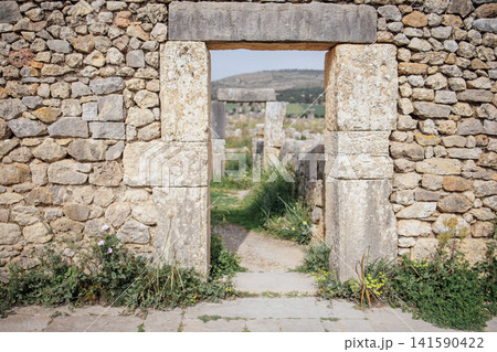 Stone archway entrance framed by weathered stone walls leading to a grassy pathway in an ancient archaeological site surrounded by hills and vegetation Stone archway entrance framed by weathered stone walls leading to a grassy pathway in an ancient archaeological site surrounded by hills and vegetation 141590422