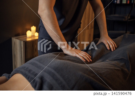 Male massage therapist providing a relaxing massage to a client covered with a dark towel in a tranquil spa environment with soft candlelight ambiance Male massage therapist providing a relaxing massage to a client covered with a dark towel in a tranquil spa environment with soft candlelight ambiance 141590476