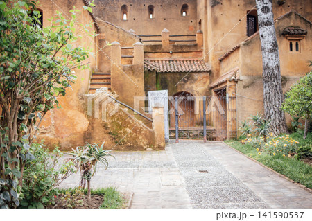 Beautiful garden pathway leads to rustic architecture with warm earthy tones, surrounded by lush greenery and vibrant flowers, creating a serene outdoor atmosphere 141590537