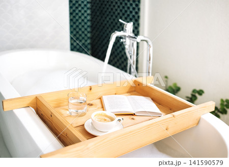 A wooden tray is attached to a bathtub filled with hot fragrant water with fluffy foam 141590579