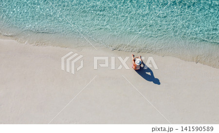 An aerial view of a young man in a white hat sitting on the ocean shore. A man enjoys the seascape. A man is relaxing on a clean sandy beach. An aerial view of a young man in a white hat sitting on the ocean shore. A man enjoys the seascape. A man is relaxing on a clean sandy beach. 141590589